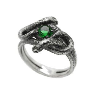 Könnte beinhalten: Ein silberner Ring mit einem grünen Edelstein in der Mitte, der mit einem detaillierten Design aus zwei verschlungenen Schlangen verziert ist.