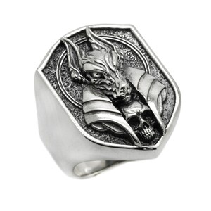 May include: A silver ring with a detailed design of a wolf's head with a skull underneath. The ring is shaped like a shield.