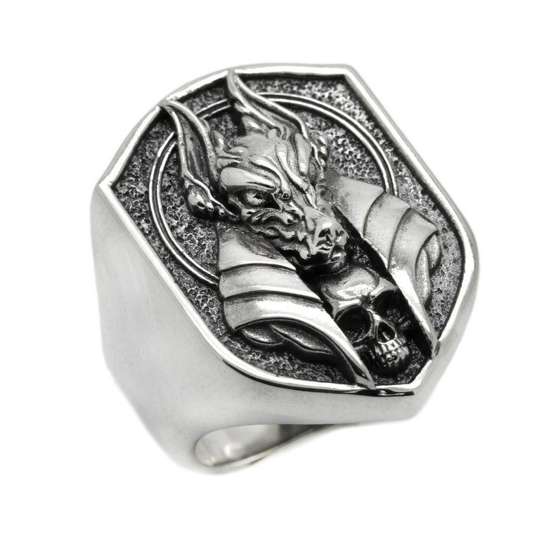 May include: A silver ring with a detailed design of a wolf's head with a skull underneath. The ring is shaped like a shield.