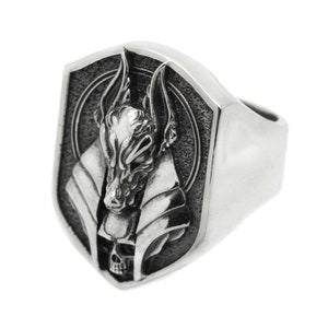 May include: A silver ring with a detailed design of an Anubis head with a skull underneath. The ring is shaped like a shield.