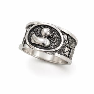 May include: A silver ring featuring a duck design within an oval frame. The ring has a textured band with additional decorative elements. The duck is in a side profile, with a detailed beak and eye. The ring is set against a white background.