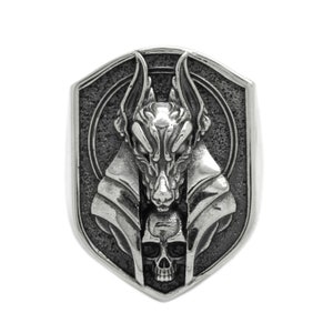 May include: A silver ring with a detailed design of an anubis head with a skull underneath. The ring is in the shape of a shield.