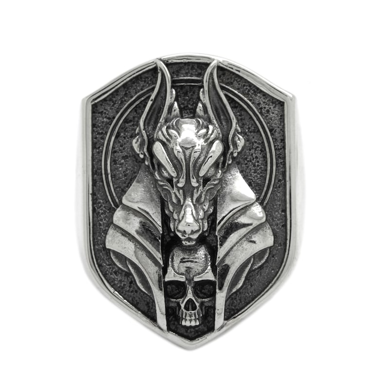 May include: A silver ring with a detailed design of an anubis head with a skull underneath. The ring is in the shape of a shield.