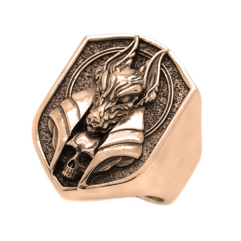 May include: A gold-toned ring with a detailed design of a wolf's head with a skull underneath. The ring is made of metal and has a textured surface.