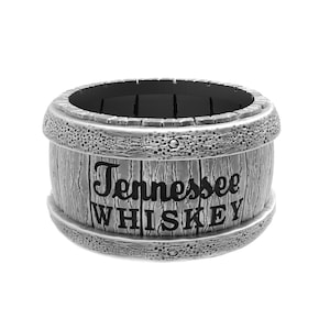 Può includere: Un contenitore a forma di botte color argento con le parole "Tennessee WHISKEY" in lettere nere. Il contenitore ha un aspetto strutturato simile al legno con fasce decorative sopra e sotto.