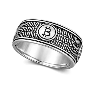 May include: Silver ring with a Bitcoin symbol in a circular design. The ring's band is engraved with binary code. The Bitcoin symbol is centered on the ring, with the binary code wrapping around the band.