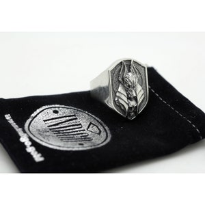 May include: A silver ring with a shield-shaped design featuring a detailed Anubis head. The ring is displayed on a black velvet pouch with a silver logo.