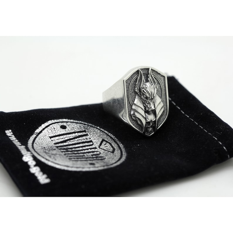 May include: A silver ring with a shield-shaped design featuring a detailed Anubis head. The ring is displayed on a black velvet pouch with a silver logo.