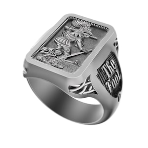 Puede incluir: Anillo plateado con un relieve detallado de un bufón con un bastón y el texto "The Fool" en el lateral. El anillo tiene una cara rectangular con un borde texturizado y detalles laterales intrincados.