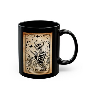 Puede incluir: Taza de cerámica negra con un diseño de carta del tarot. La carta representa un esqueleto leyendo un libro, con el texto "THE READER" en la parte inferior. El diseño incluye una mariposa, flores y fases lunares.