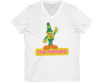 Puede incluir: Camiseta blanca con cuello en V con un personaje de botella de dibujos animados con detalles en verde y amarillo, zapatos naranjas y un dedo levantado. El texto "THE MOTHER" está en un rectángulo amarillo.