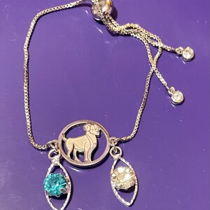 May include: A silver adjustable bracelet with a silver charm featuring the zodiac symbol for Aries. The charm is attached to two dangling charms, one with a blue gemstone and the other with a clear gemstone.