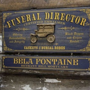 K&ouml;nnte beinhalten: Ein Schild aus Holz im Vintage-Stil f&uuml;r einen Bestatter. Das Schild ist blau lackiert mit goldenen Buchstaben und zeigt eine Illustration eines schwarzen und goldenen Leichenwagens. Der Text auf dem Schild lautet "FUNERAL DIRECTOR CERTIFIED EMBALMER Offering Understanding and Integrity COFFINS CASKETS & BURIAL ROBES Black Drapes and Crepes Reutrd BELA FONTAINE BETHANY HILL MORTUARY".