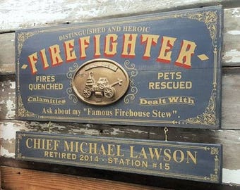 Può includere: Un cartello in legno blu con lettere dorate che recita "Distinguished and Heroic Firefighter." Il cartello presenta un emblema di camion dei pompieri in metallo dorato e il testo "Fires Quenched, Calamities Dealt With, Pets Rescued." Il cartello include anche il testo "Ask about my "Famous Firehouse Stew."." Sotto il cartello c'è un cartello in legno blu più piccolo con lettere dorate che recita "Chief Michael Lawson Retired 2014 - Station #15."