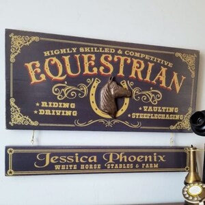 May include: A dark blue wooden sign with gold lettering that reads "HIGHLY SKILLED & COMPETITIVE EQUESTRIAN *RIDING *DRIVING *VAULTING *STEEPLECHASING". A brown horse head sculpture is attached to the sign. A smaller dark blue wooden sign with gold lettering reads "Jessica Phoenix WHITE HORSE STABLES & FARM".