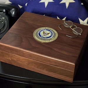 May include: A rectangular dark brown wooden box featuring a circular gold and blue United States Navy emblem on the lid. The emblem is inscribed with the text COLIN RICHARD MASSEY and COMMISSIONED MAY 2026. A pair of wire-rimmed glasses rests nearby.
