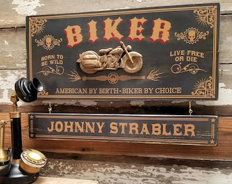 May include: Black wooden sign with gold lettering that reads "BIKER" with a gold motorcycle graphic and the text "BORN TO BE WILD" and "LIVE FREE OR DIE". Below the main sign is a smaller black wooden sign with gold lettering that reads "JOHNNY STRABLER".