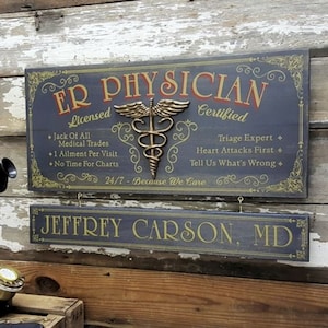 May include: A dark blue wooden sign with gold lettering and a gold medical symbol. The sign reads "ER Physician Licensed Certified" and includes the text "Jack of All Medical Trades + 1 Ailment Per Visit + No Time For Charts" and "Triage Expert + Heart Attacks First + Tell Us What's Wrong". The sign also reads "24/7 - Because We Care". A smaller dark blue wooden sign with gold lettering reads "JEFFREY CARSON, MD".