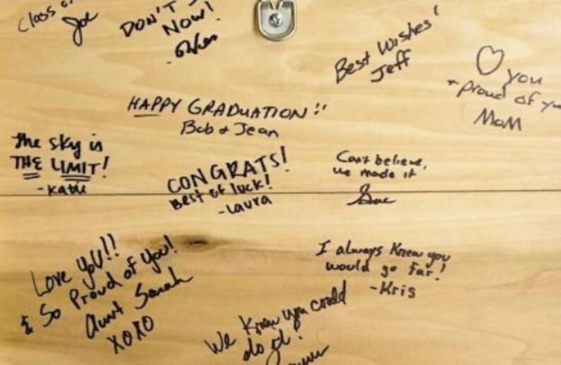 May include: A wooden sign with handwritten messages of congratulations and well wishes. The sign reads "Happy Graduation!" "The sky is the limit!" "Congrats! Best of luck!" "Love you!! & So proud of you! Aunt Sarah XOXO" "Best Wishes Jeff" "Can't believe we made it!" "I always knew you would go far!" "You - proud of you Mom"