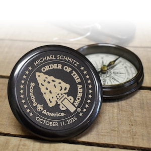 May include: A black compass with a gold inscription that reads "Michael Schmitz Order of the Arrow Scouting America October 11, 2025".