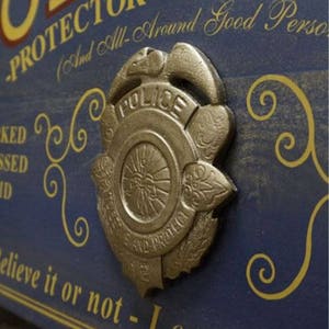 May include: A blue wooden sign with gold lettering and a gold metal police badge. The sign reads "Protector of the Innocent (And All-Around Good Person)".