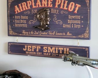May include: A dark purple wooden sign with gold text reading AIRPLANE PILOT FOR HIRE, featuring a bronze-colored pilot head relief. Below, a smaller sign reads JEFF SMITH WINGS EST. MAY 25TH 1981. Includes a model airplane and leather goggles.