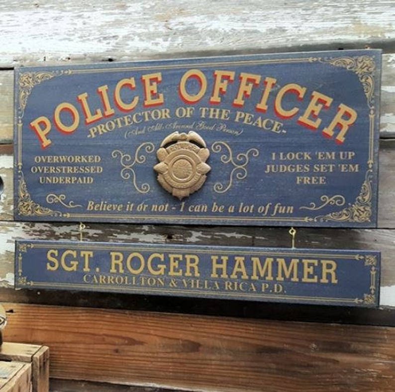 May include: A blue wooden sign with gold lettering that reads "Police Officer - Protector of the Peace" with a gold badge and the text "Overworked, Overstressed, Underpaid" and "I lock 'em up, Judges set 'em free". Below the sign is a smaller blue wooden sign with gold lettering that reads "Sgt. Roger Hammer, Carrollton & Villa Rica P.D."