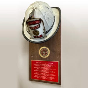 May include: A white firefighter helmet with a red and gold badge that reads "ASST CHIEF ANTON FD" is mounted on a wooden plaque. The plaque has a gold medallion with a fire department logo and a red plaque with the "A Firefighter's Prayer" text.