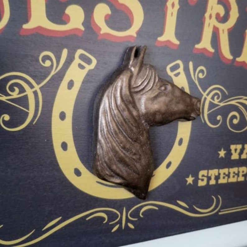 May include: A rustic wooden sign with a gold horseshoe and a bronze horse head sculpture. The sign reads "Oldest Ride * Ya'll Steeple Chase*"