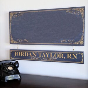 K&ouml;nnte beinhalten: Zwei dunkelblaue Holzschilder mit goldenen Buchstaben. Das obere Schild hat einen dekorativen goldenen Rand. Das untere Schild lautet "JORDAN TAYLOR, RN EMORY UNIVERSITY".