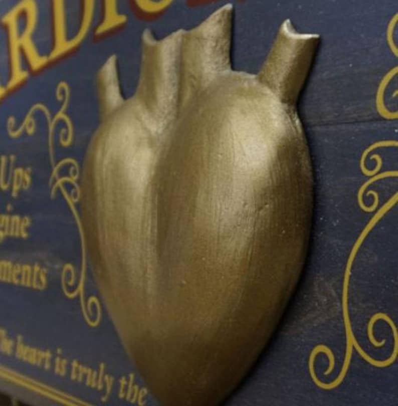 K&ouml;nnte beinhalten: Ein goldfarbenes herzf&ouml;rmiges Ornament mit einer strukturierten Oberfl&auml;che. Das Herz ist an einem dunkelblauen Holzuntergrund befestigt, auf dem in goldenen Lettern steht: "Das Herz ist wirklich der Motor aller H&ouml;hen und Tiefen".