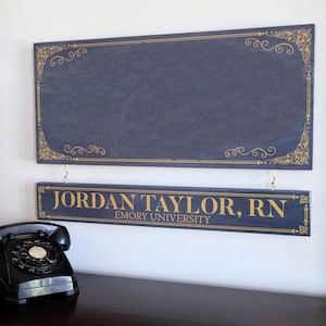 K&ouml;nnte beinhalten: Zwei dunkelblaue Holzschilder mit goldenen Buchstaben. Das obere Schild hat einen dekorativen goldenen Rand. Das untere Schild lautet "JORDAN TAYLOR, RN EMORY UNIVERSITY".