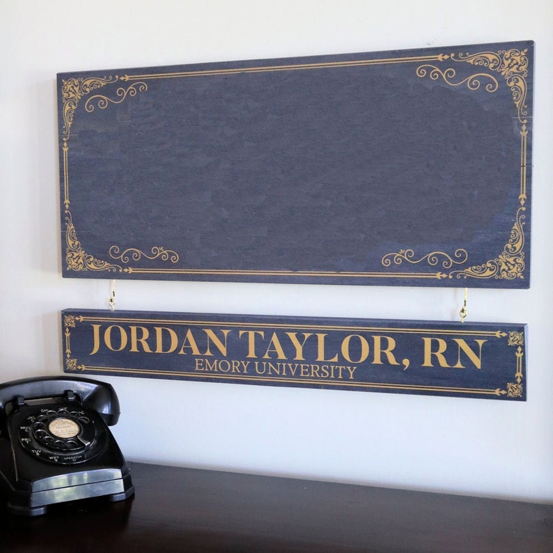 K&ouml;nnte beinhalten: Zwei dunkelblaue Holzschilder mit goldenen Buchstaben. Das obere Schild hat einen dekorativen goldenen Rand. Das untere Schild lautet "JORDAN TAYLOR, RN EMORY UNIVERSITY".