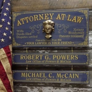 K&ouml;nnte beinhalten: Drei Holzschilder mit goldenen Buchstaben auf blauem Hintergrund. Das oberste Schild lautet "ATTORNEY AT LAW" mit einer goldenen B&uuml;ste einer Augenbinden tragenden Frau in der Mitte. Das mittlere Schild lautet "ROBERT G. POWERS Law Office of Powers & McCain". Das unterste Schild lautet "MICHAEL C. MCCAIN Law Office of Powers & McCain".