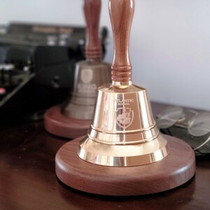 May include: Two brass school bells with wooden handles and bases. The bells have the words "ATLANTIC SCHOOL" and a crest engraved on them. The bells are sitting on a dark wooden surface.
