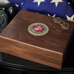 May include: A rectangular dark brown wooden box featuring a circular gold and blue United States Marine Corps emblem on the lid. The emblem includes the text COLIN RICHARD MASSEY and COMMISSIONED MAY 2026. A pair of wire-rimmed glasses rests on the lid.