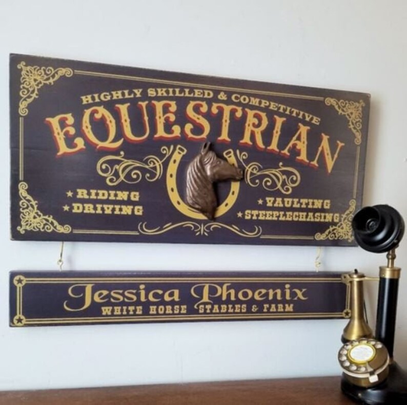May include: A dark blue wooden sign with gold lettering that reads "HIGHLY SKILLED & COMPETITIVE EQUESTRIAN *RIDING *DRIVING *VAULTING *STEEPLECHASING". A brown horse head sculpture is attached to the sign. A smaller dark blue wooden sign with gold lettering reads "Jessica Phoenix WHITE HORSE STABLES & FARM".