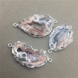 May include: Three natural agate stone pendants with silver-colored metal edging and attached loops. The stones feature a mix of gray, white, brown, and purple hues. These pendants are ideal for jewelry making.