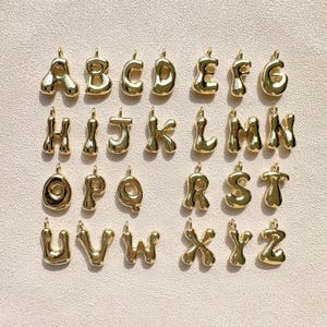 Puede incluir: Una colección de colgantes de alfabeto dorados, cada uno con un diseño redondeado en forma de burbuja. Se muestran las letras de la A a la Z, cada una con un pequeño lazo en la parte superior para sujetar a una cadena o pulsera. Los colgantes están dispuestos sobre una superficie beige claro.