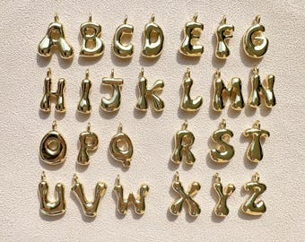 May include: A collection of gold-colored alphabet pendants, each with a rounded, bubble-like design. The letters A through Z are displayed, each with a small loop at the top for attaching to a chain or bracelet. The pendants are arranged on a light beige surface.