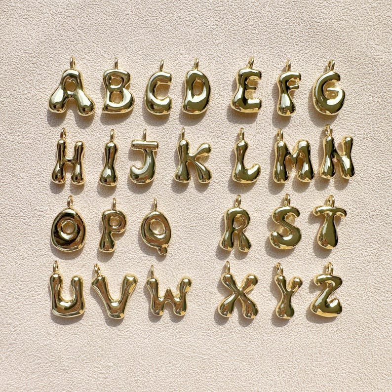 May include: A collection of gold-colored alphabet pendants, each with a rounded, bubble-like design. The letters A through Z are displayed, each with a small loop at the top for attaching to a chain or bracelet. The pendants are arranged on a light beige surface.