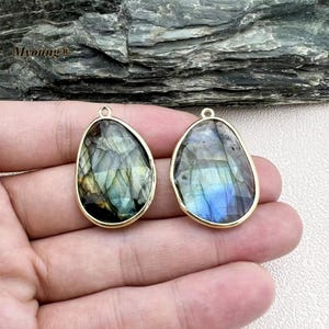 Puede incluir: Dos colgantes de tono dorado con una piedra preciosa de labradorita en forma de lágrima. Las piedras tienen un brillo iridiscente azul, verde y gris.