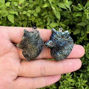 Può includere: Due ciondoli in labradorite scolpita a forma di teste di lupo. I ciondoli hanno un bordo in metallo color argento e un anello per una catena. Le teste di lupo sono dettagliate con pelliccia e lineamenti del viso e mostrano sfumature iridescenti blu e verdi.