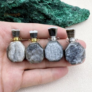 May include: Four small, faceted gemstone bottles with dark gray and white crystal formations. Each bottle has a silver or gold-colored metal collar and a dark gray stopper. The bottles are held in a hand, with a green stone in the background.