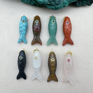 Puede incluir: Ocho colgantes de pez tallados en varios colores, incluyendo turquesa, rojo, verde y negro. Cada colgante tiene una fianza dorada y un ojo de piedra preciosa. Los peces se exhiben sobre una superficie de color claro.