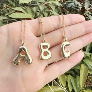 May include: Three gold-colored bubble letter necklaces spelling "ABC". Each pendant is a different letter, suspended from a delicate gold chain. The necklaces are displayed against a background of green foliage.