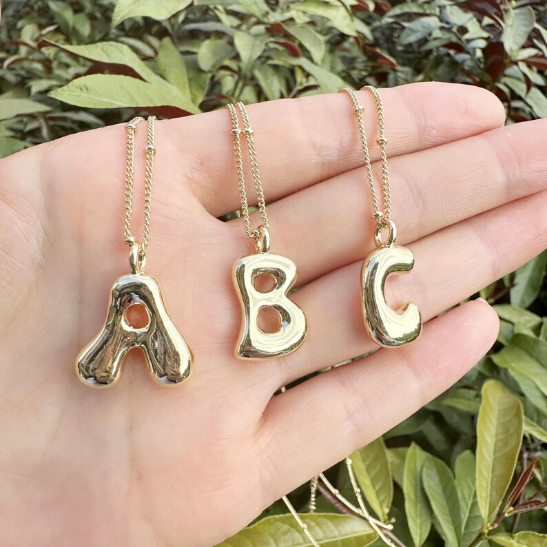 May include: Three gold-colored bubble letter necklaces spelling "ABC". Each pendant is a different letter, suspended from a delicate gold chain. The necklaces are displayed against a background of green foliage.