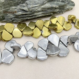 May include: Close-up of metallic beads in gold and silver tones. The beads have an irregular, organic shape, resembling small pebbles. The beads are arranged in rows, with a natural stone slab in the background. The Myoung logo is visible.