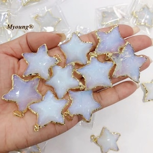 May include: A collection of white opal star-shaped pendants with gold-toned edges. The pendants are arranged in a hand, with some of the pendants overlapping.