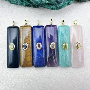 Op de afbeelding: Zes rechthoekige edelsteenhangers in verschillende kleuren: zwart, bruin, blauw, paars, turquoise en roze. Elke hanger heeft een gouden beugel en een kleine, ovaalvormige edelsteen in het midden. Gepresenteerd tegen een witte achtergrond.